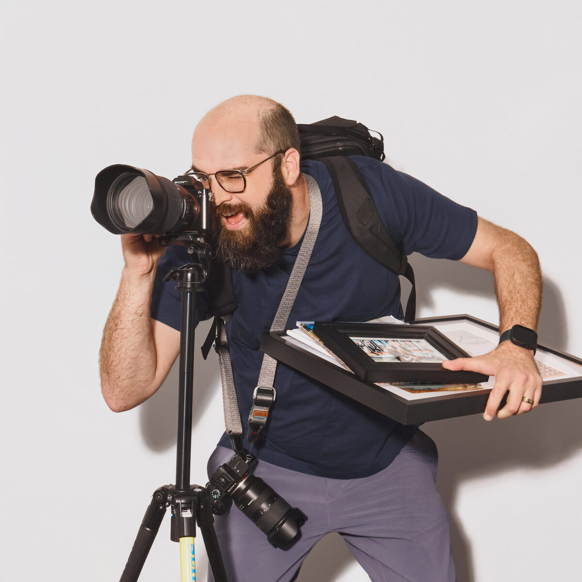 Jake Muller photographing with a DSLR on a tripod, carrying framed prints and gear