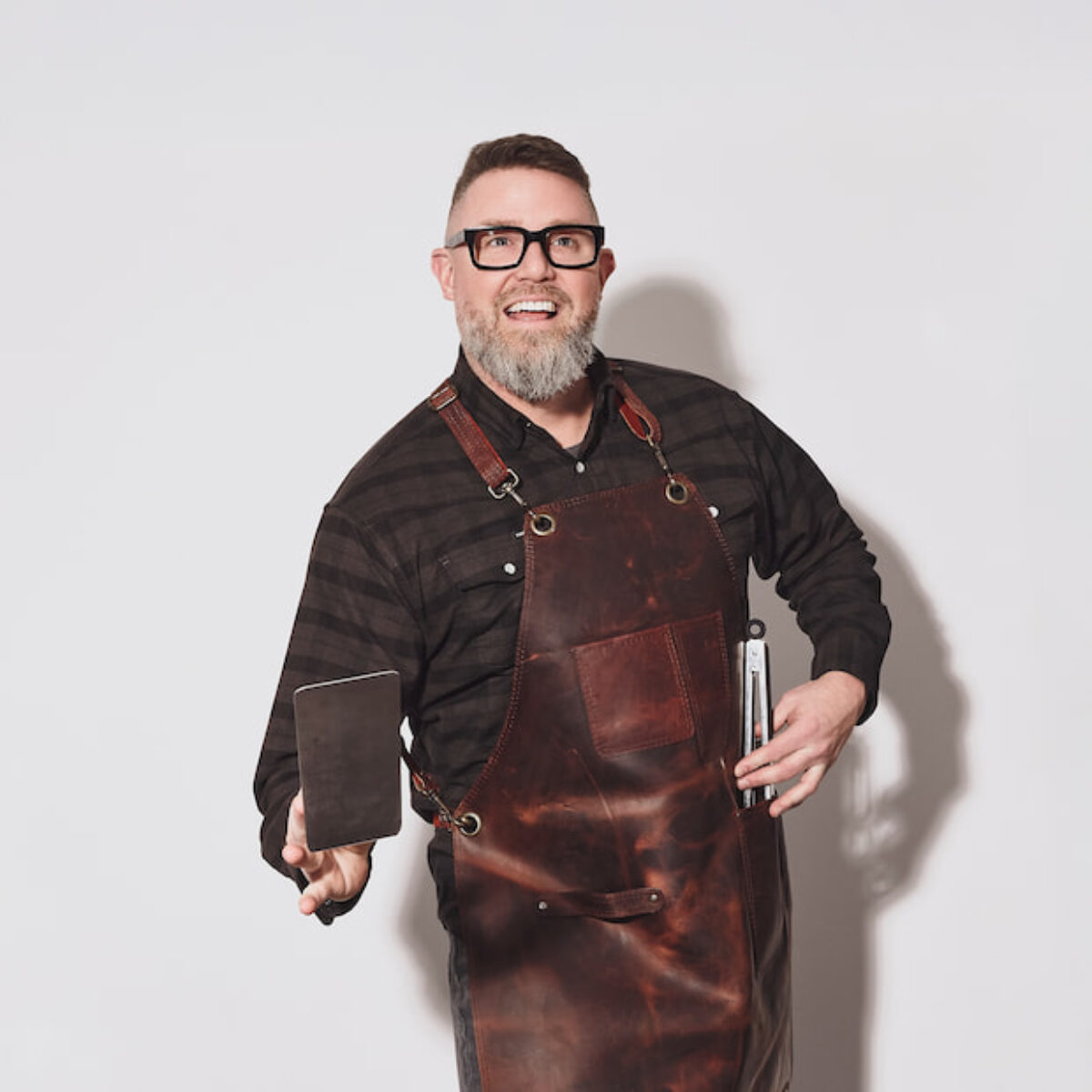 Ryan Howell grilling in a leather apron, holding a spatula and tongs